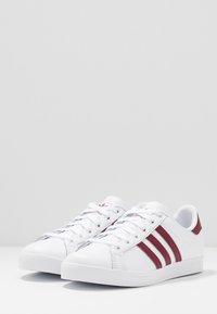 Vita sneakers i syntetmaterial, med tre röda ränder, platta snören och rundad tåkonstruktion. Slät yta, låg profil.