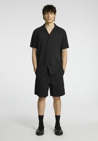 Camicia nera a maniche corte con bottoni e vestibilità rilassata, abbinata a pantaloni corti neri, indossata con calzini neri e scarpe, design minimale.