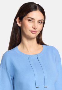 Jasnoniebieski sweter w dzianinę z okrągłym dekoltem, z dwoma ozdobnymi sznurkami i teksturowanym wykończeniem w okolicach ramion.