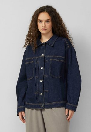 Jonge vrouw met krullend haar, gekleed in een donkerblauwe spijkerjas met grote voorzakken en lichtgekleurde broek.