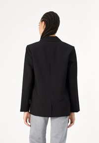 Črn prilagojen blazer z enim razporkom, z žepkami in dolgimi rokavi, izdelan iz gladkega materiala. Kombiniran z sivimi hlačami.