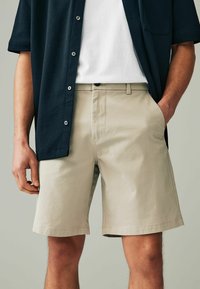 Beige Shorts mit geradem Schnitt, aus einem Baumwollmix gefertigt. Verfügt über Seitentaschen und einen Knopfverschluss, kombiniert mit einem dunklen Hemd.