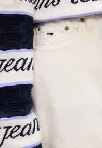 Gros plan de la poche d’un pantalon en velours côtelé blanc avec petit logo drapeau Tommy Hilfiger et manche de pull tricoté bleu marine et blanc avec texte.