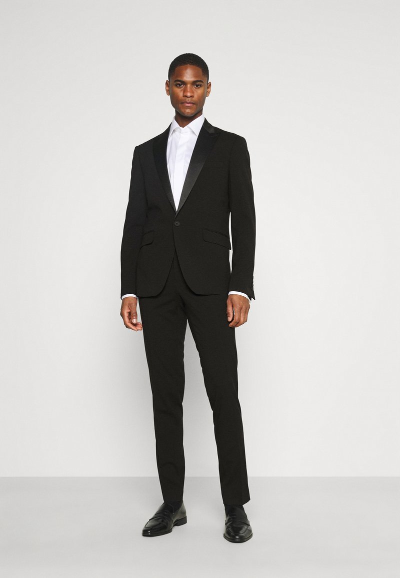 Costum negru cu revere din satin, cămașă albă, pantaloni slim fit și pantofi negri.  Material neted cu un design croit.