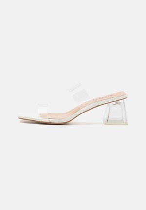 Hvid åben tå mule-sandal med klare dobbelte stropper og en transparent geometrisk blokhæl på hvid baggrund.