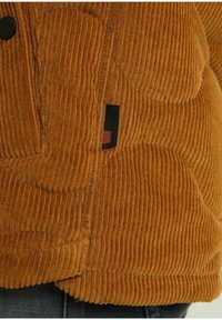 Gros plan sur une veste en velours côtelé marron avec une étiquette en tissu noire et marron près de la couture latérale et un bouton noir visible.