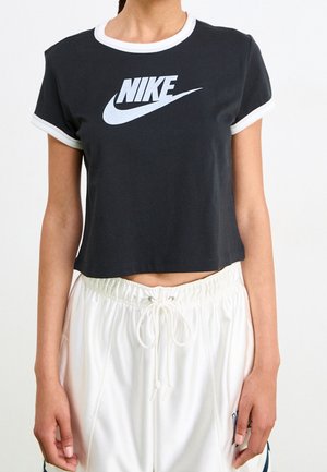 Kobieta ubrana w czarny krótki t-shirt Nike z białym wykończeniem i luźne białe spodnie ze sznurkiem na jednolitym tle.