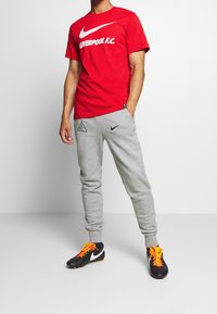 Camiseta roja de Nike con estampado de "LIVERPOOL F.C.", combinada con pantalones de chándal grises con detalles y logo en negro. Zapatillas negras con cordones naranjas.