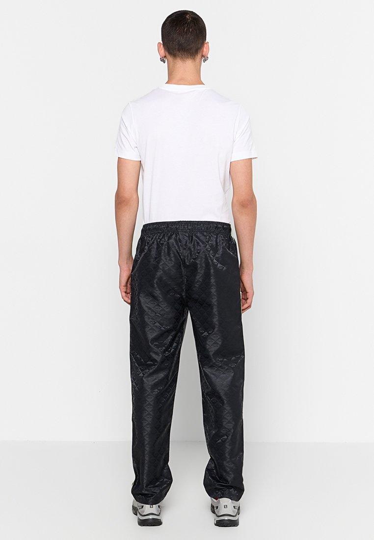 Persona in piedi di spalle che indossa una maglietta bianca, pantaloni neri a fantasia e sneakers grigie, davanti a uno sfondo bianco uniforme.