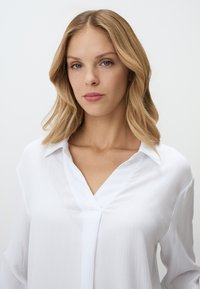 Blouse blanche à col en V, avec une texture subtile. Manches longues et coupe décontractée. Design uni sans motifs ni accents visibles.