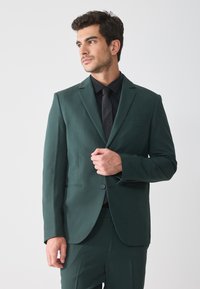 Dunkelgrüne Anzugjacke mit tailliertem Schnitt, Ein-Knopf-Verschluss, Reverskragen und glatter Textur. Trägt ein schwarzes Hemd und eine Krawatte.