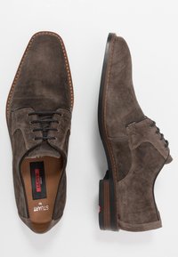 Chaussures de ville en daim marron avec un bout arrondi, présentant un laçage noir et une semelle en caoutchouc marron contrastante.