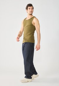 Débardeur côtelé olive avec un col large, associé à un jean droite bleu foncé. Modèle portant des baskets crème, se tenant de profil.