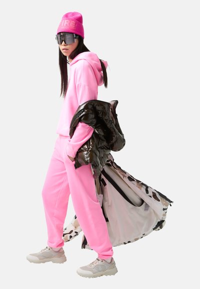 Helle pinke Kapuzenjacke und Jogginghosen, glänzende schwarze Pufferjacke, graue klobige Sneakers und eine rosa Strickmütze mit Logo. Dunkle Sonnenbrille.