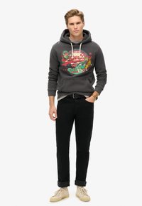 Sudadera gris con estampado gráfico colorido, jeans negros y zapatillas beige. Cuenta con un bolsillo tipo canguro y capucha con cordón. Conjunto casual.