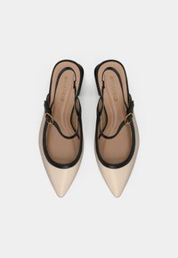 Sandales à talons beiges et noires avec un design à bout pointu, présentant une bordure noire et un détail de boucle dorée sur la bride de cheville.