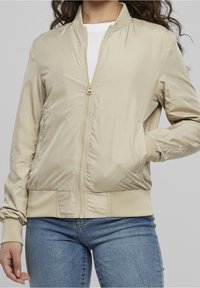 Kvinna i en beige zip-up bomberjacka med ribbad muddar och fåll, matchad med blå jeans, står med en hand i fickan.