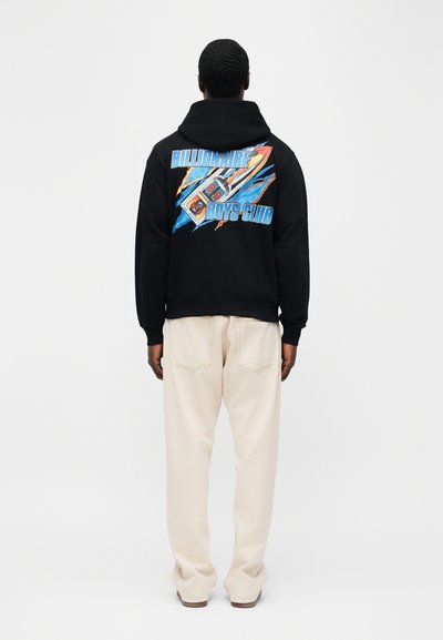 Άνδρας που φοράει μαύρο φούτερ με κουκούλα με πολύχρωμο σχέδιο πυραύλου και το κείμενο "Billionaire Boys Club" στην πλάτη, συνδυασμένο με ανοιχτό μπεζ παντελόνι.