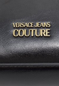 Surface en cuir noir texturé avec un texte métallique doré affichant "VERSACE JEANS COUTURE" centré dessus.
