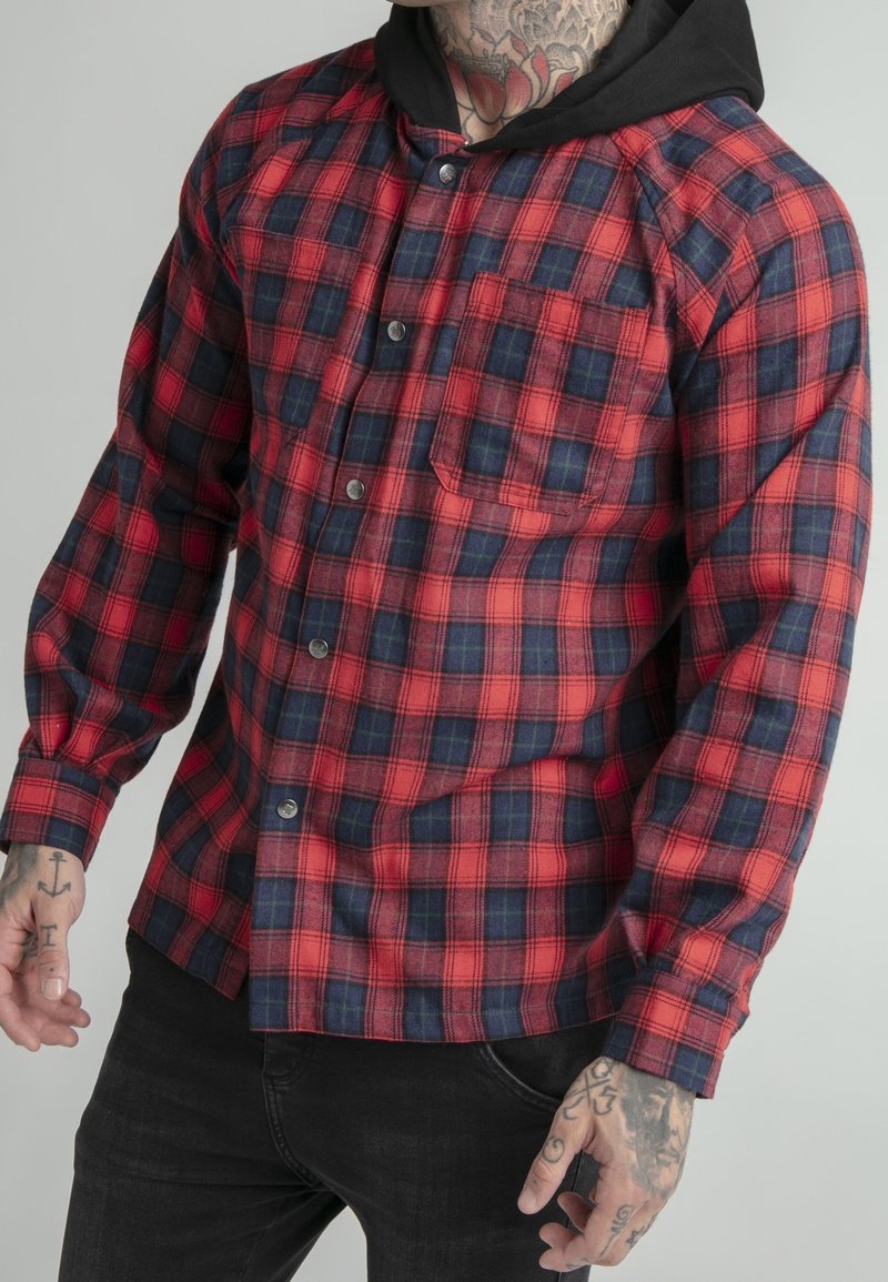Homme portant une chemise à carreaux rouge et bleu marine sur un sweat à capuche noir, montrant des mains et un cou tatoués, sur un fond uni.