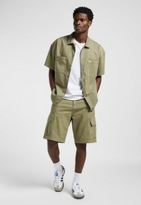 Chemise olive à manches courtes et short cargo assorti, avec des poches poitrine doubles, portée sur un t-shirt blanc, et des baskets décontractées.