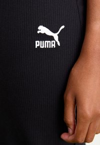 Nahaufnahme einer Hand, die neben einem schwarzen, gerippten Stoff ruht, auf dem ein weißes, gesticktes Puma-Logo mit springender Katze und Text zu sehen ist.