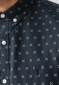 Marineblauwe short-sleeve shirt met witte kruispatronen, knoopsluiting vooraan, en een klein geborduurd detail aan de zijkant. Soepele stoftextuur.