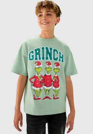 Vihreä t-paita, jossa on kolme Grinch-hahmoa punaisissa juhlapuvuissa, joissa on valkoista reunusta. Rohkea "GRINCH" -teksti turkoosina kuvan yläpuolella.