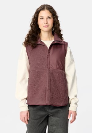 Donna con capelli ricci che indossa un gilet in pile color granata sopra una camicia chiara a maniche lunghe e pantaloni scuri su uno sfondo semplice.