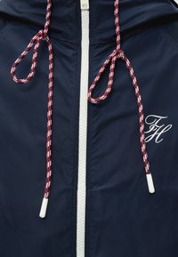 Veste marine avec fermeture éclair blanche, cordons rouges et blancs attachés en boucles, et initiales cursives blanches "FH" sur la poitrine.