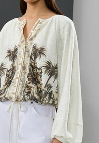 Blouse blanche avec un imprimé floral de palmier en marron et beige, dotée d'un décolleté en trou de serrure et de détails brodés sur le devant.