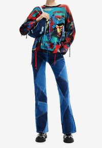 Pull multicolore avec des motifs abstraits, manches longues et détails à franges, associé à un jean bleu en patchwork à pattes d'éléphant et des bottes noires chunky.