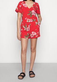 Romper floreale rosso con maniche corte, scollo a V, vita elastica e tasche. Presenta motivi floreali rosa e verdi. Indossato con sandali neri.