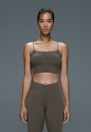 Femme debout face à l’objectif, portant un crop top marron moulant avec des fines bretelles et des leggings marron taille haute sur fond gris.