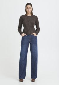 Kobieta stoi z rękami w kieszeniach, ubrana w brązowy kardigan z guzikami w warkoczowy wzór, szerokie niebieskie dżinsy i brązowe buty na białym tle.