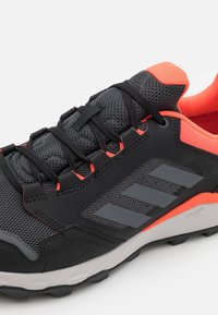 Scarpa sportiva nera con tomaia in mesh, brillanti accenti arancioni, stringhe spesse, strisce laterali grigie e suola in gomma testurizzata per una buona aderenza.