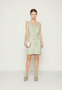 Vestido sem mangas de tom verde claro com bordados florais, cintura ajustada e decote em V; combinado com sapatos de plataforma a condizer e uma clutch.