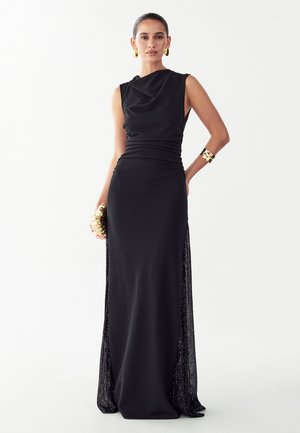 Femme portant une robe noire longue sans manches avec un décolleté drapé, tenant une pochette dorée et portant un bracelet et des boucles d'oreilles en or.