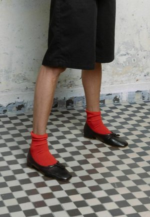 Ballerine in pelle nera con punta quadrata e dettaglio a fiocco, abbinate a calze rosse a costine, indossate con pantaloni corti neri su un pavimento piastrellato decorato.