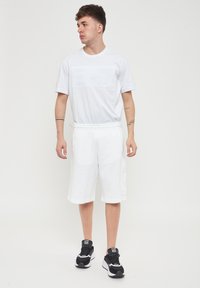 T-shirt bianco in cotone con logo "EA7" in rilievo, abbinato a pantaloni corti bianchi con tasche laterali e sneakers sportive nere.