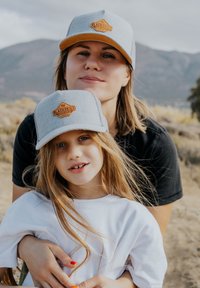 Szürke snapback sapkák barna bőrön készült "BLACKSKIES" felirattal. A háttérben lágy, elmosódott hegyek és sivatagi növényzet található.