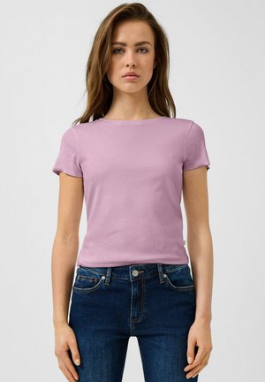 Młoda kobieta stojąca, ubrana w jasnofioletowy top z krótkim rękawem i ciemnoniebieskie jeansy na jednolitym jasnym tle.