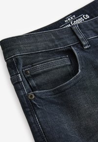 Jean en denim bleu foncé avec une poche avant pour pièce de monnaie montrant des rivets et des coutures visibles près de la taille et des passants de ceinture.
