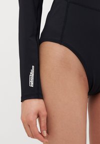 Fato de banho de manga longa preto com design ajustado, costuras planas e um rótulo de proteção UV na manga. Tecido suave e elástico.