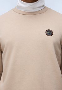 Sudadera beige hecha de tejido texturizado, con un parche redondo del logo en la parte izquierda del pecho con detalles en negro y naranja.