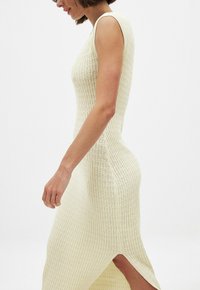 Strickkleid ohne Ärmel in Cremefarbe mit geribbelter Textur, verfügt über einen seitlichen Schlitz und eine taillierte Passform, die die Körperkonturen betont.