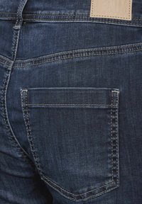 Pantalones vaqueros de mezclilla azul oscuro con un bolsillo trasero con costuras en contraste y una etiqueta de cuero cerca de la cintura, presentando una textura suave.