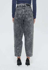 Personne portant un jean gris délavé taille haute avec des poches arrière et des chaussures à talons hauts noires, se tenant devant un fond uni.