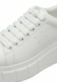 Vit sneakers med en texturerad syntetisk ovandel, utrustad med sex tygöglor, platta snören och en tjock, ruggad sula. Små ventilationshål på sidan.