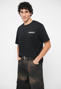 Jovem com cabelo encaracolado veste uma t-shirt preta da Double A, cinto preto com ilhoses e calças cargo pretas com manchas de tintura castanha, com as mãos nos bolsos.